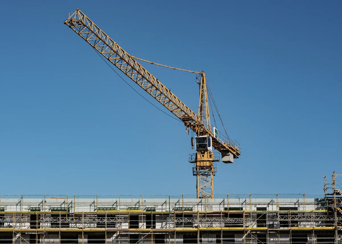 Großer gelber Turmdrehkran ragt vor einem klaren blauen Himmel über eine im Bau befindliche Gebäudestruktur mit Gerüsten und Absperrungen.