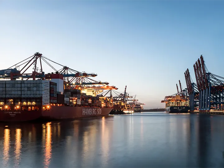 Abendliche Aufnahme des Hamburger Hafens mit mehreren großen Containerschiffen und Containerbrücken. Beleuchtete Hafenanlagen und Schiffe spiegeln sich im ruhigen Wasser.